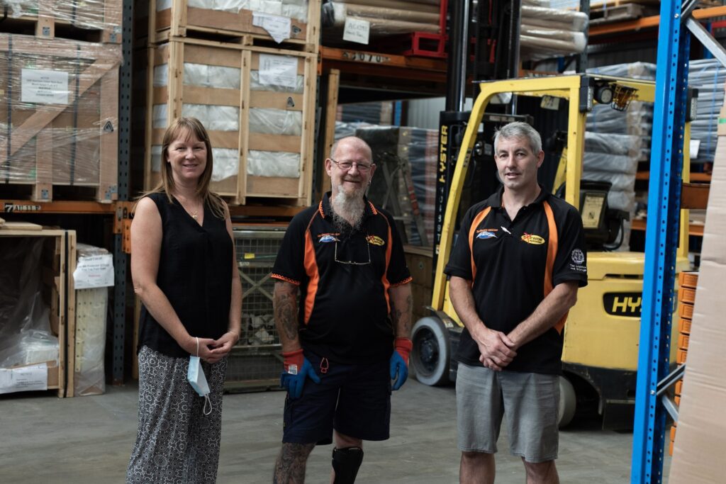 Two middle-aged men and a middle-aged woman standing in a warehouse.