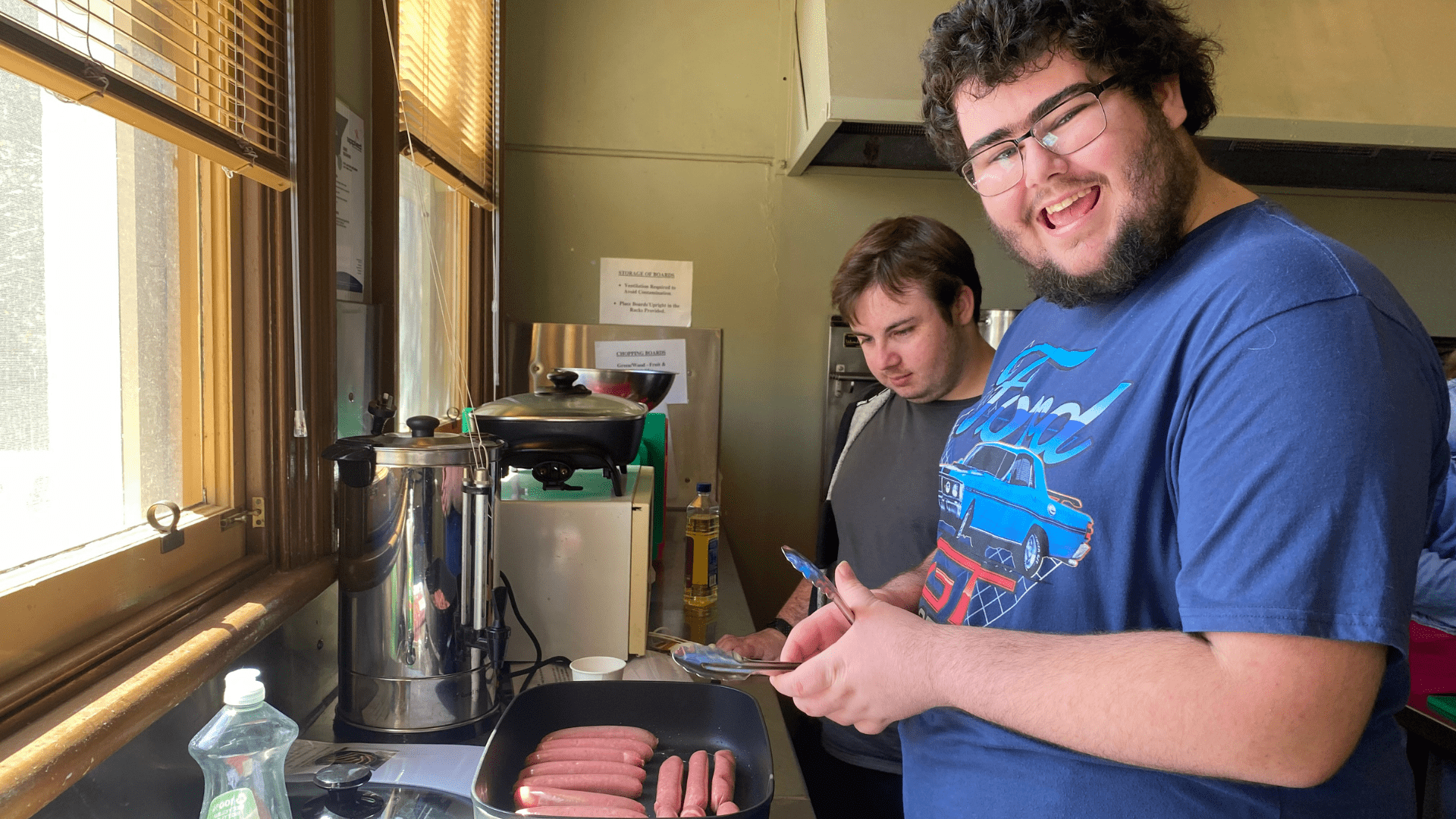 Young men happily cooking sausages in a kitchen.