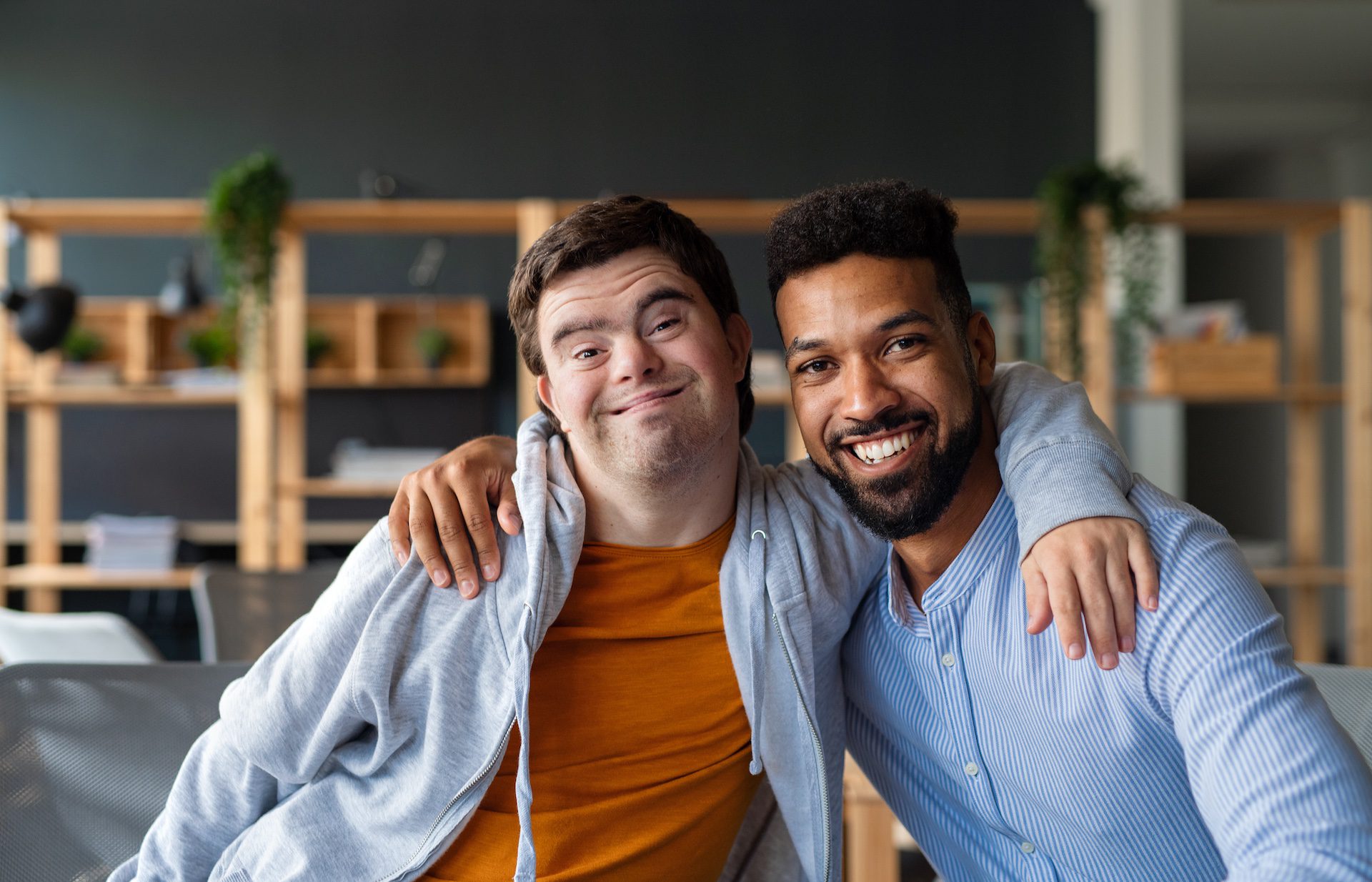 Young man with Down syndrome and his tutor, arms around each other, smiling at the camera indoors at school.