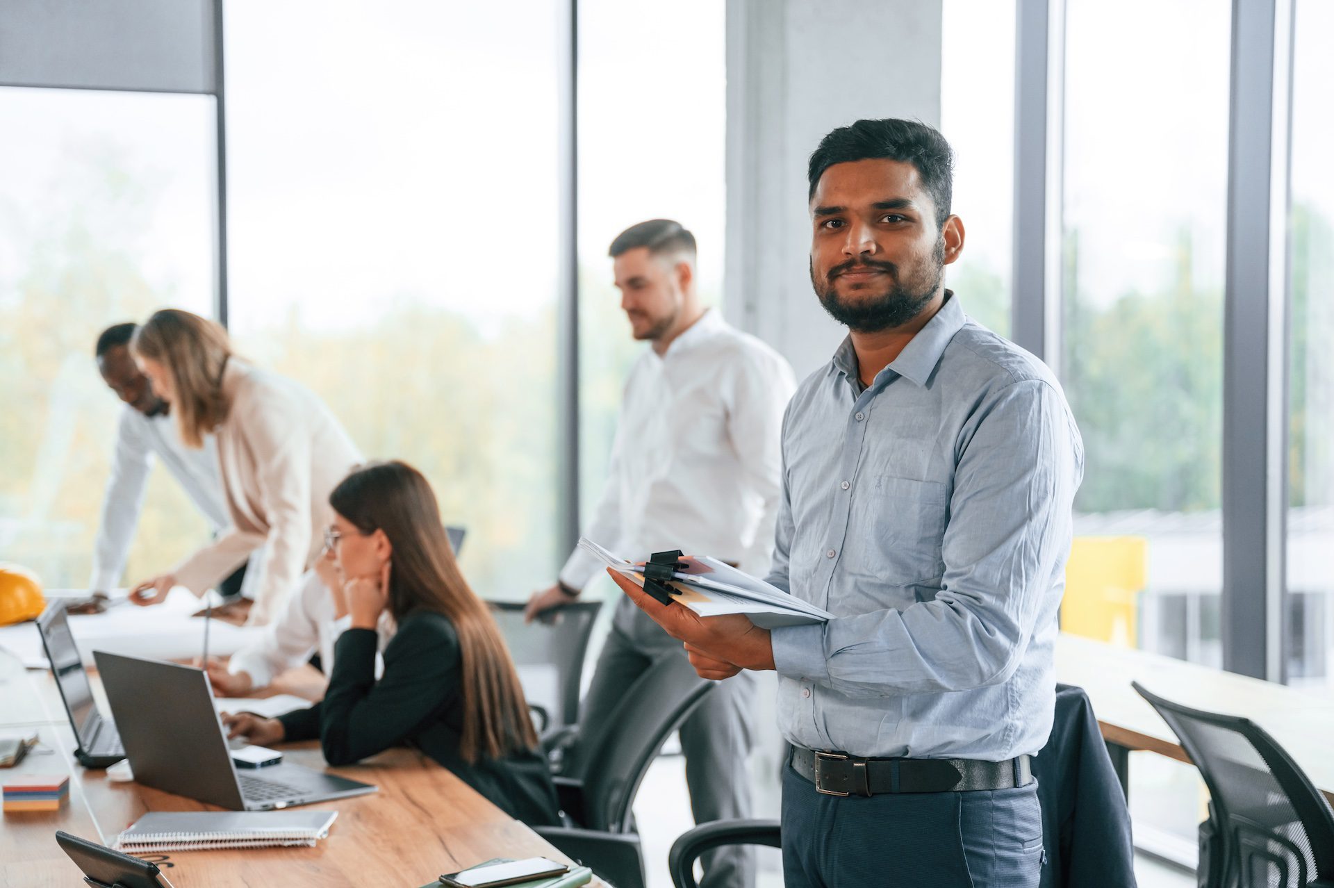 Indian man standing in front of colleagues using laptops, with a group of office workers collaborating indoors.