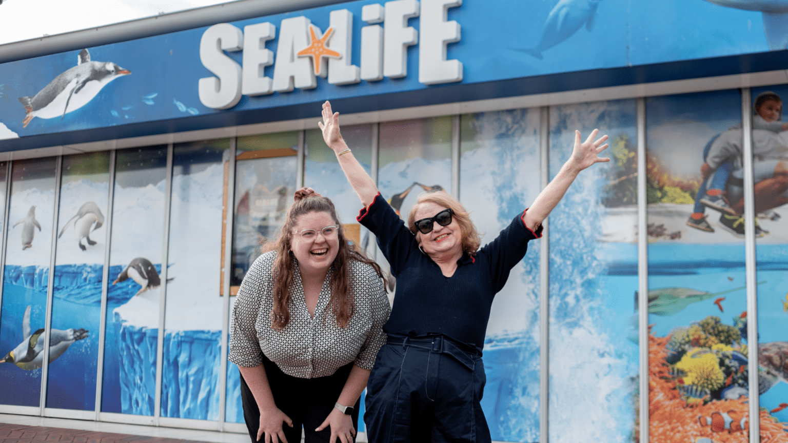 Bianca is in a dark blue blouse with her hands in the air and Gemma is on her left bending over in laughter