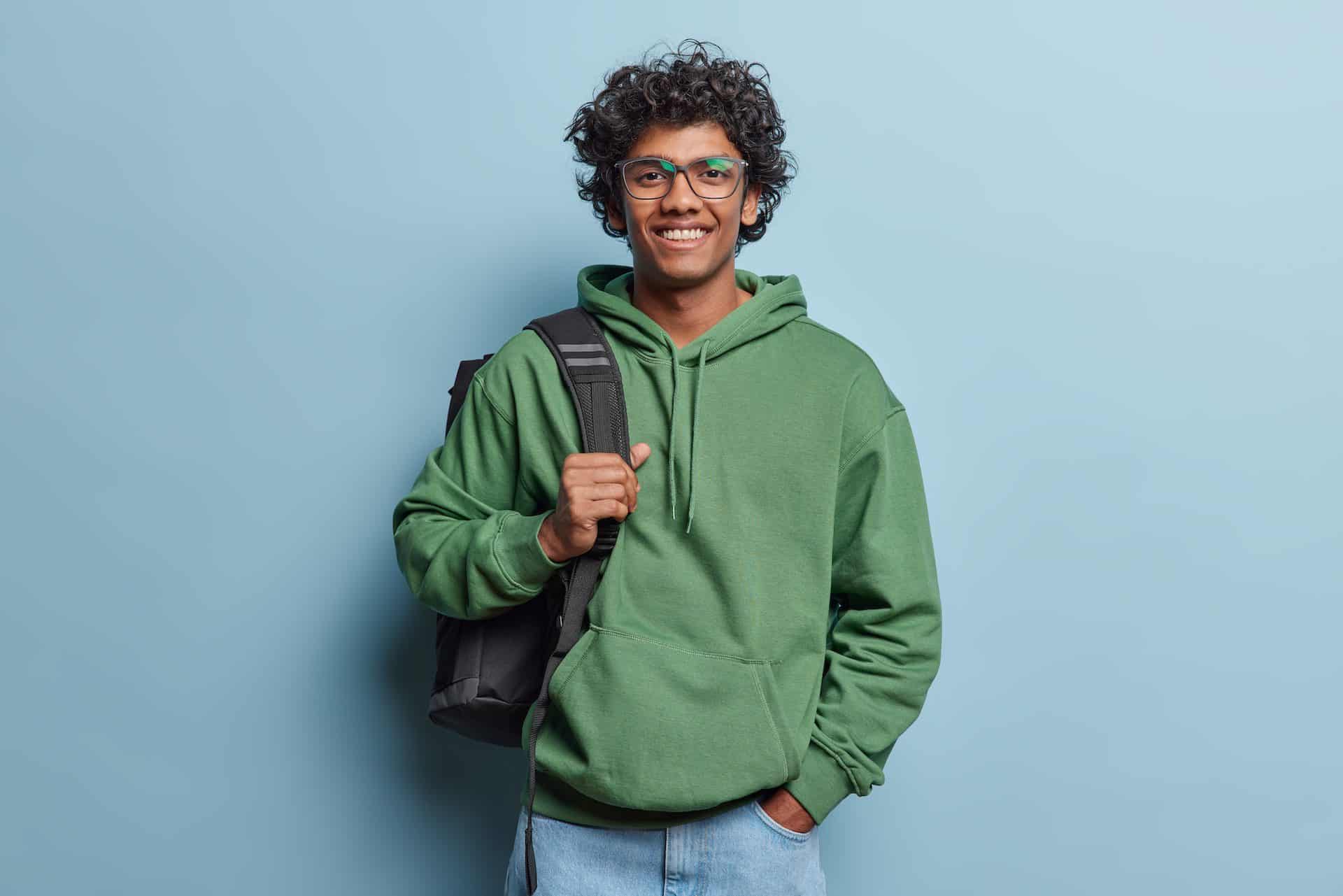 Happy young Hindu male student in a green hoodie, jeans, and a black bag, smiling in a studio against a blue background.