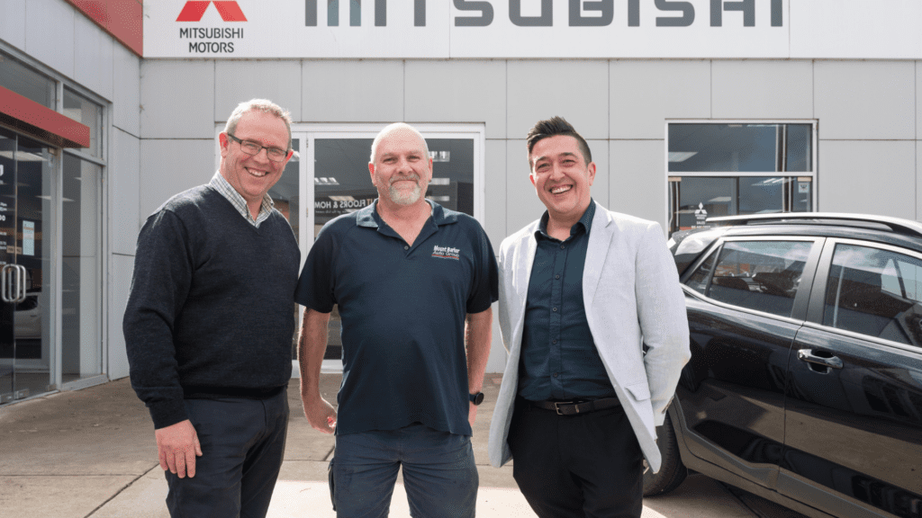 Three smiling men standing together in front of a car dealership.