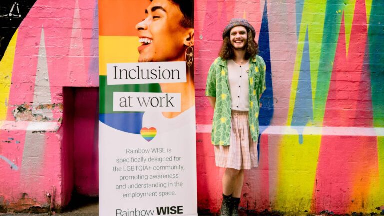 Noa standing next to Rainbow WISE signage, in front of a colourful mural