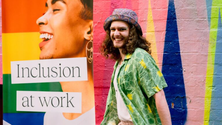 Noa standing next to Rainbow WISE signage, in front of a colourful mural