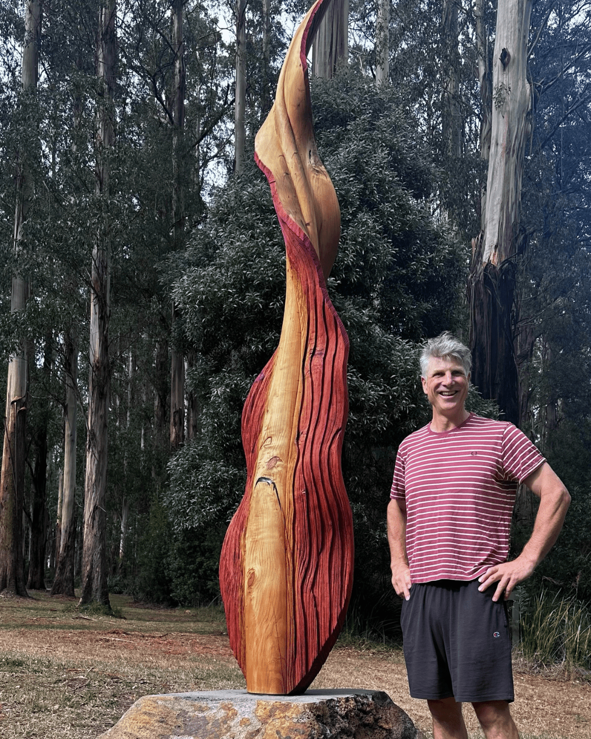 Anton standing outdoors with his hands on his hips, next to a large wooden sculpture he created