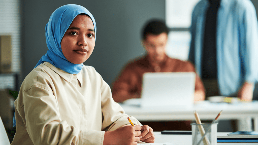 Young lady wearing a hijab and holding a pencil with office workers in the background
