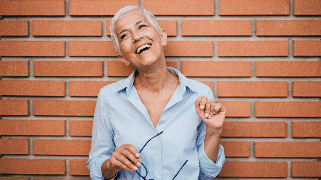 A woman with white hair, leaning against a red brick wall.