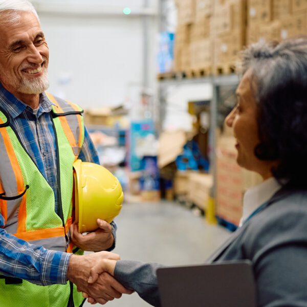 Two mature workers smiling and shaking hands — one in a high-vis vest holding a yellow hard hat, the other in a grey blazer