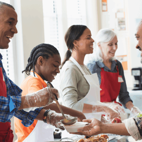 A diverse group of people smiling together, with some serving food from behind a table and another person receiving a meal.