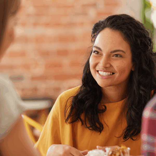 A woman in a mustard-yellow top smiles warmly while talking with two other people in a casual indoor setting