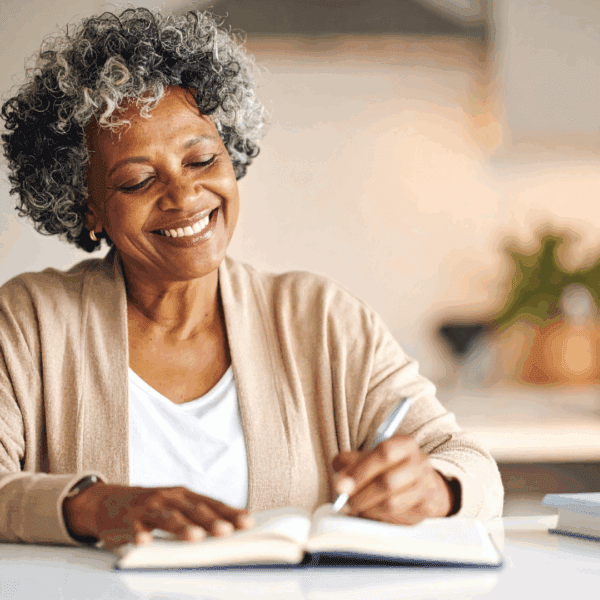 An older woman smiling as she writes in a notebook at a kitchen table with a cup of coffee beside her