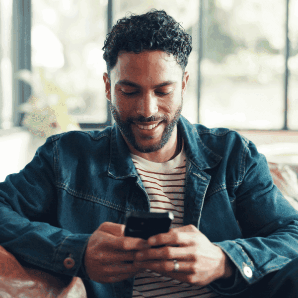 Man sitting on a couch smiling while looking at his phone, wearing a denim jacket and striped shirt in a bright living room