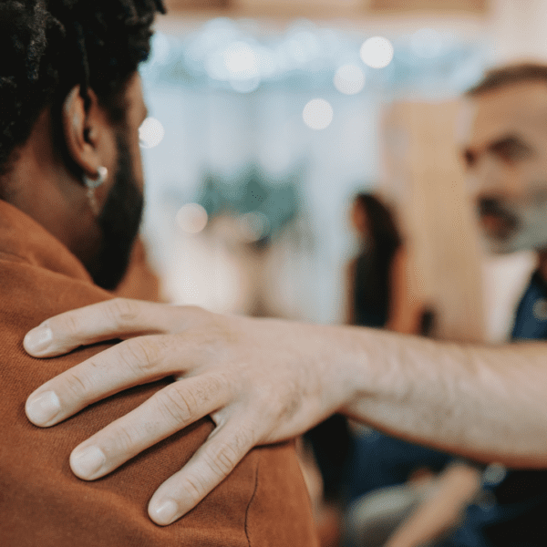 One person places a supportive hand on another person’s shoulder during a workplace conversation.