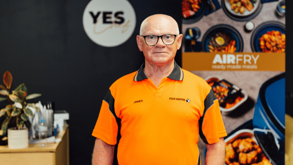 An older man with glasses and a bald head stands in front of a 'YES' sign and a poster advertising 'AIRFRY ready made meals.' He wears an orange polo shirt with 'JAMES' and 'FUJI XEROX' embroidered on it. The background includes a plant and a display of food items in bowls.