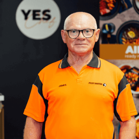An older man with glasses and a bald head stands in front of a 'YES' sign and a poster advertising 'AIRFRY ready made meals.' He wears an orange polo shirt with 'JAMES' and 'FUJI XEROX' embroidered on it. The background includes a plant and a display of food items in bowls.
