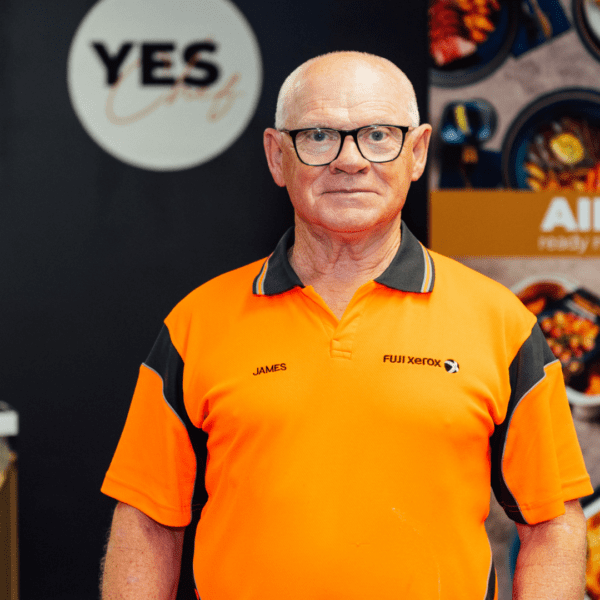 An older man with glasses and a bald head stands in front of a 'YES' sign and a poster advertising 'AIRFRY ready made meals.' He wears an orange polo shirt with 'JAMES' and 'FUJI XEROX' embroidered on it. The background includes a plant and a display of food items in bowls.