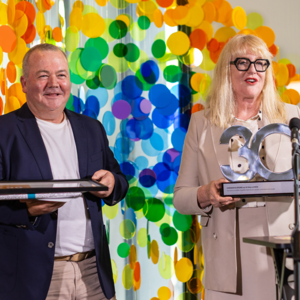 Man wearing white t-shirt under navy blazer holding a framed certificate. He is stood next to a mature blonde woman in a cream coat holding a silver '30' trophy