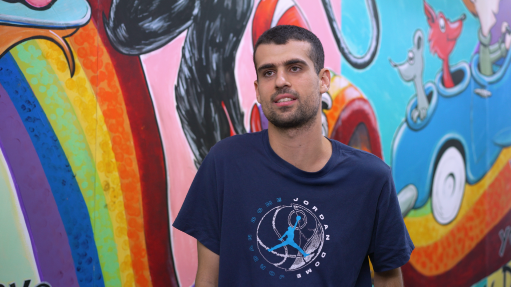 A man wearing a navy T-shirt standing in front of a bright mural with a rainbow and playful cartoon characters.