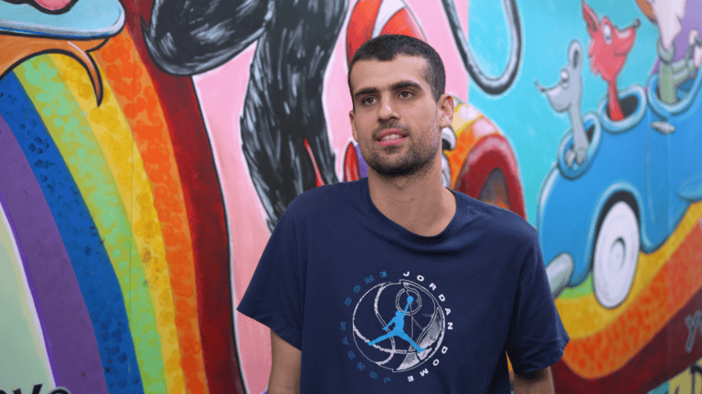 A man wearing a navy T-shirt standing in front of a bright mural with a rainbow and playful cartoon characters.
