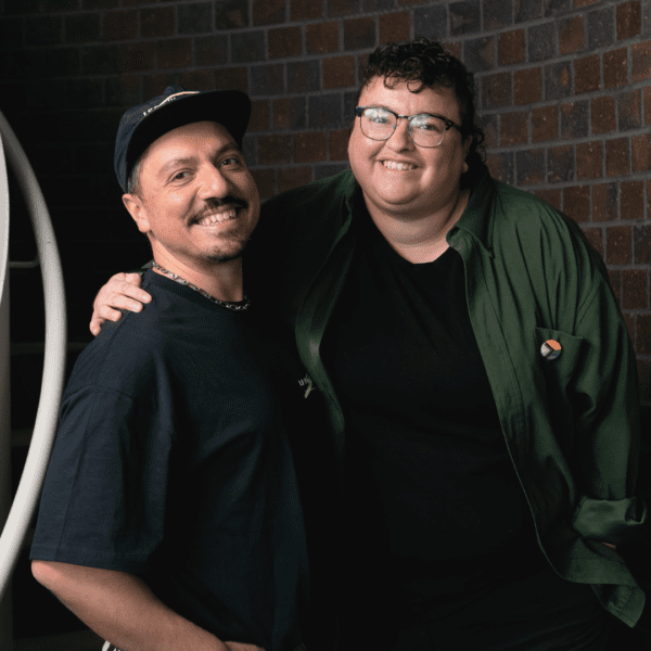 Two people smiling and standing together with their arms around each other beside a brick wall.