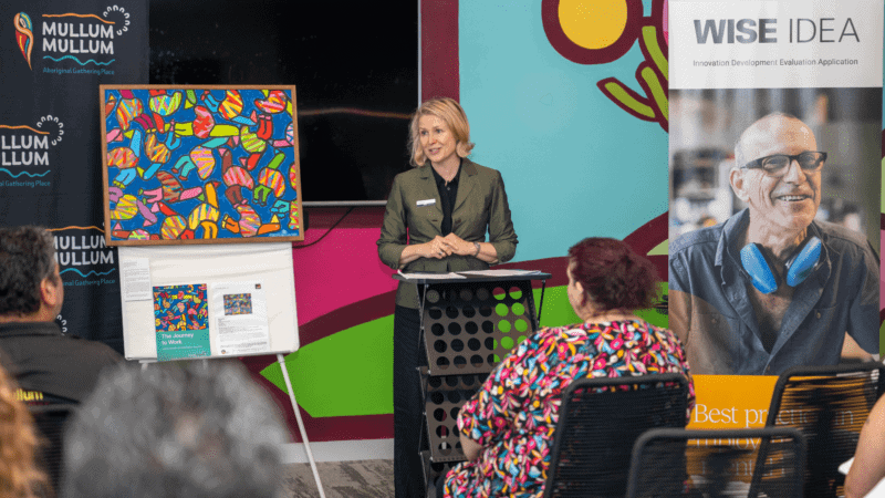 A speaker at a podium during an event, with banners for 'Mullum Mullum Aboriginal Gathering Place' and 'WISE IDEA,' and a colorful abstract painting on display.