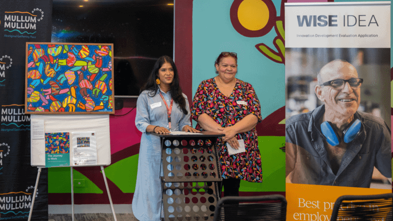 Two women presenting at a podium, flanked by banners for 'Mullum Mullum Aboriginal Gathering Place' and 'WISE IDEA,' with a vibrant abstract painting on an easel