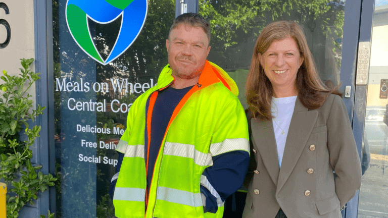 Man wearing a hi-vis jacket over a black t-shirt standing smiling next to a women wearing a grey blazer over a white top and black pants