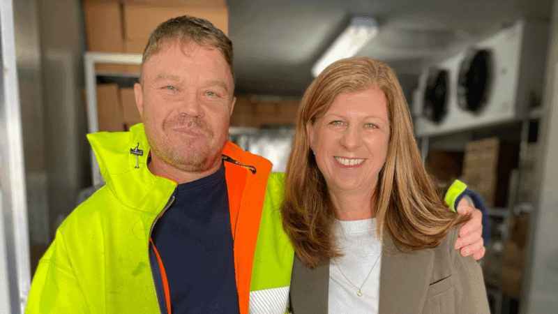 Close up photo of man wearing a hi-vis jacket over a black t-shirt standing smiling next to a women wearing a grey blazer over a white top
