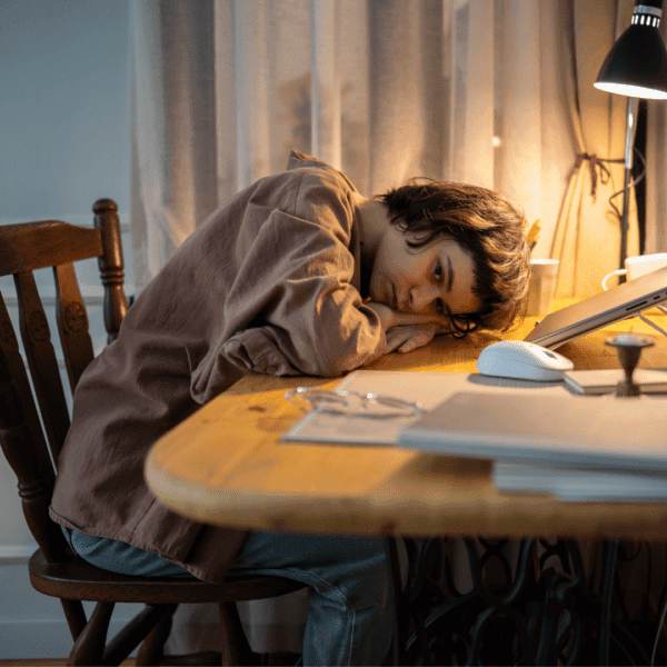 Young person resting their head on a desk beside a laptop, appearing tired or unmotivated while studying or working from home