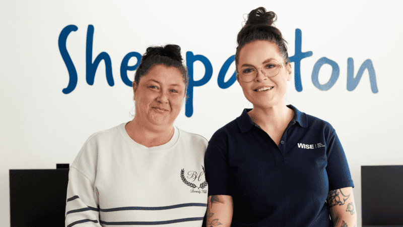 Two women stand smiling in front of a wall with the word &ldquo;Shepparton.&rdquo;