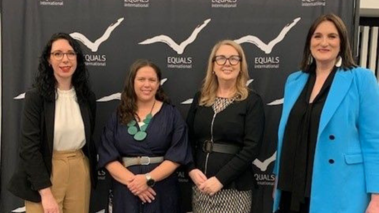Four women standing in a line, smiling at the camera, in front of a black background with a white bird pattern