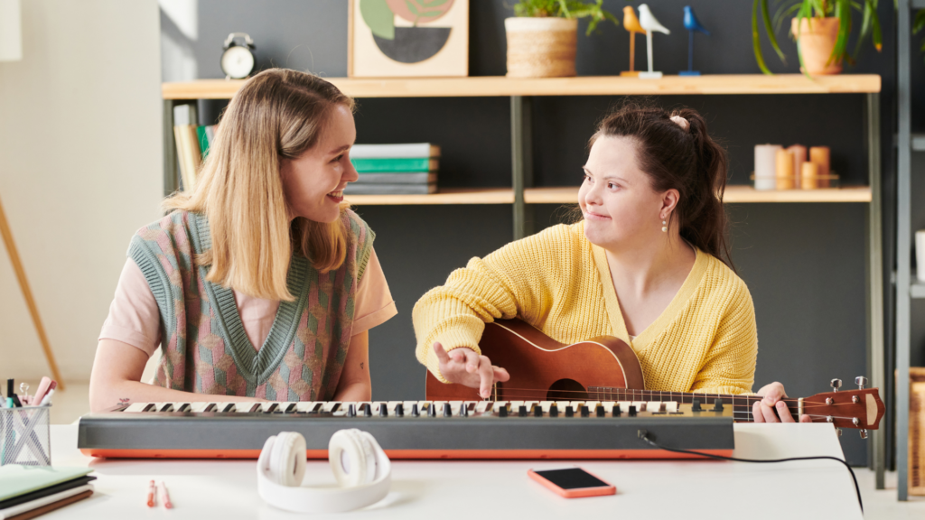 Two women sitting at a desk with a keyboard on it. The woman on the right wears a yellow jumper while holding a guitar and the woman on the left wears a grey vest. They are both looking at eachother, smiling.