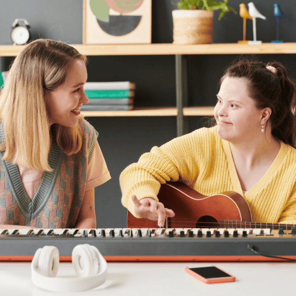 Two women sitting at a desk with a keyboard on it. The woman on the right wears a yellow jumper while holding a guitar and the woman on the left wears a grey vest. They are both looking at eachother, smiling.