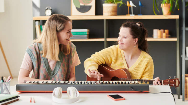 Two women sitting at a desk with a keyboard on it. The woman on the right wears a yellow jumper while holding a guitar and the woman on the left wears a grey vest. They are both looking at eachother, smiling.