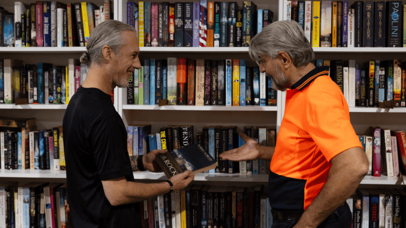 Patrick in a fluorescent orange shirt, standing next to a man in a black t-shirt, in front of a book shelf filled with books