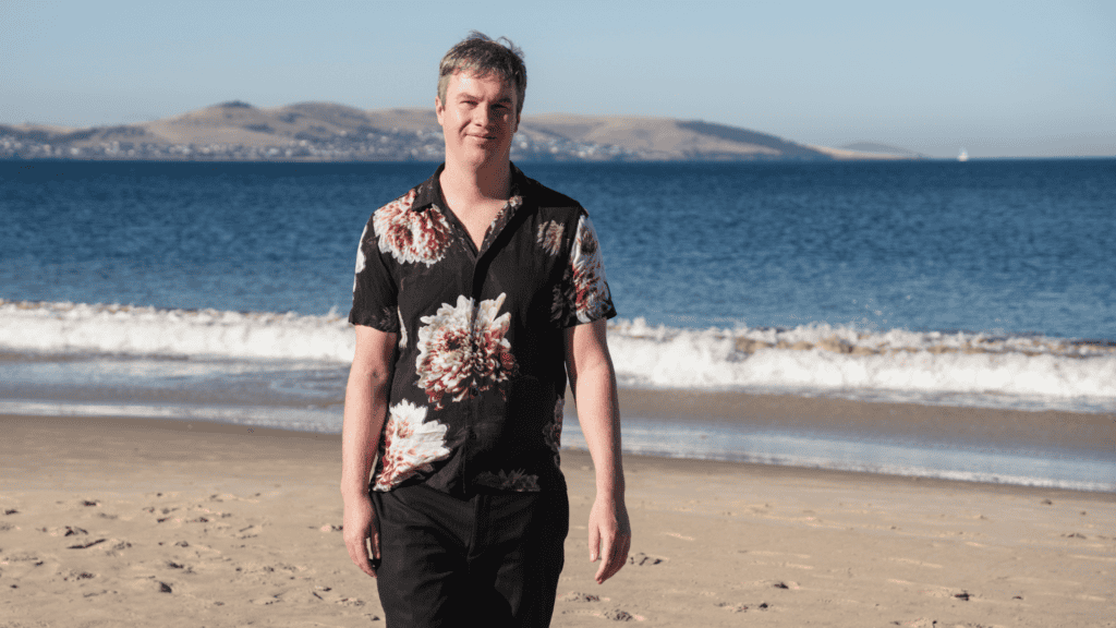 Riley, walking on the beach, wearing a black buttoned shirt with florals on it.