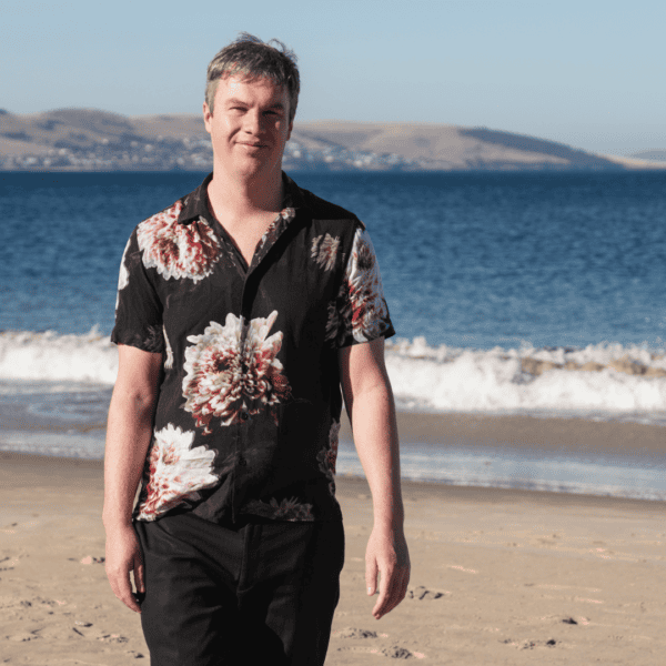 Riley, walking on the beach, wearing a black buttoned shirt with florals on it.
