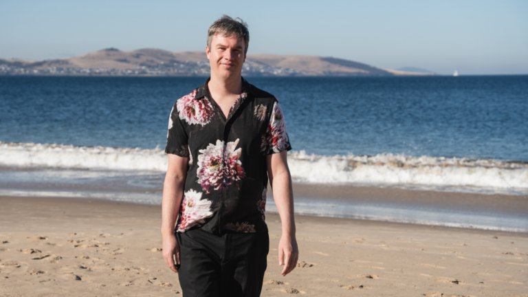 Riley, walking on the beach, wearing a black buttoned shirt with florals on it.