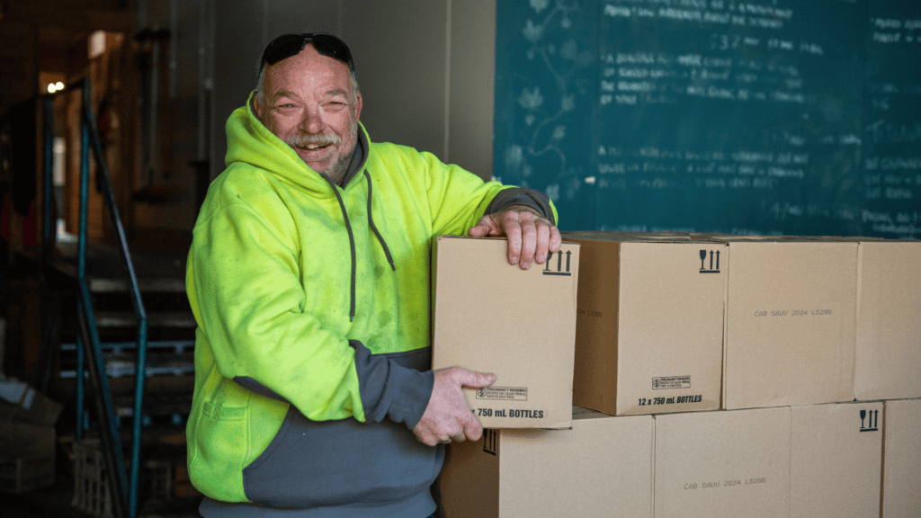Smiling man in a hi-vis hoodie holding a wine box beside stacked cases in a winery