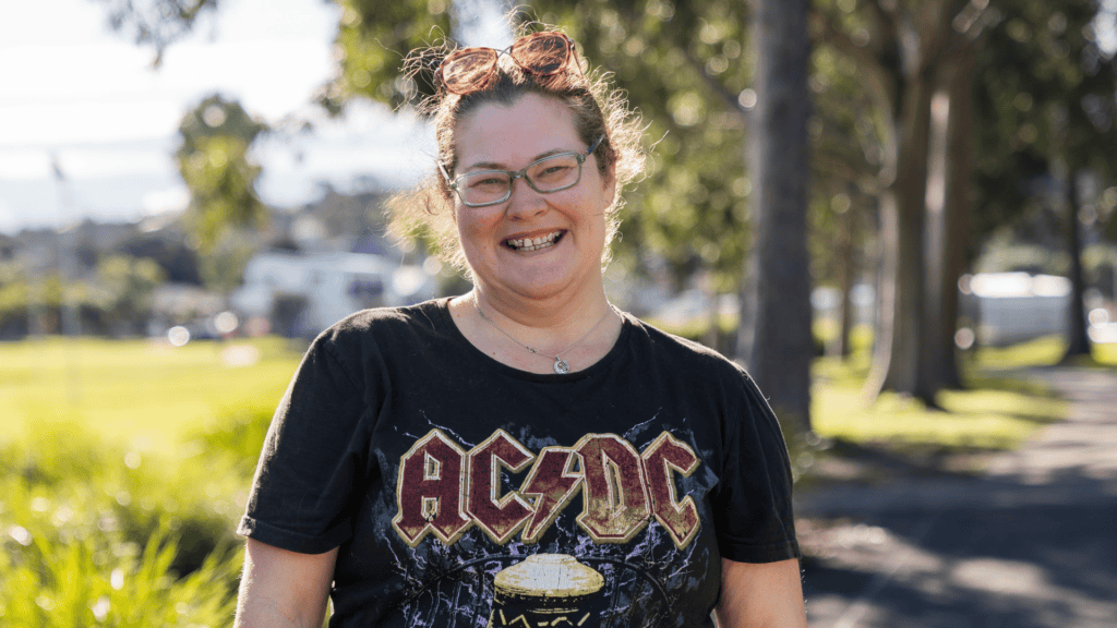 A smiling woman with brown hair and green-framed glasses stands in a sunny park, wearing a black AC/DC t-shirt.