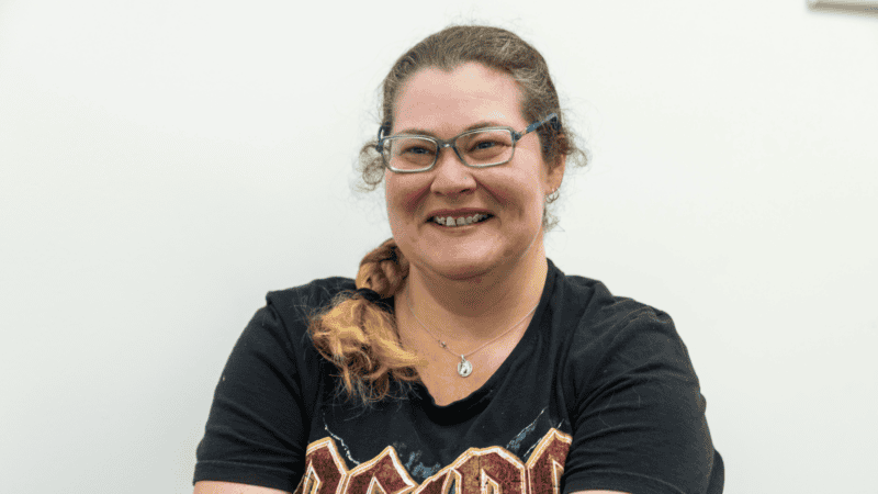 Close-up of a smiling woman with her light brown hair in a braid, wearing teal-framed glasses and a black AC/DC t-shirt against a plain background.