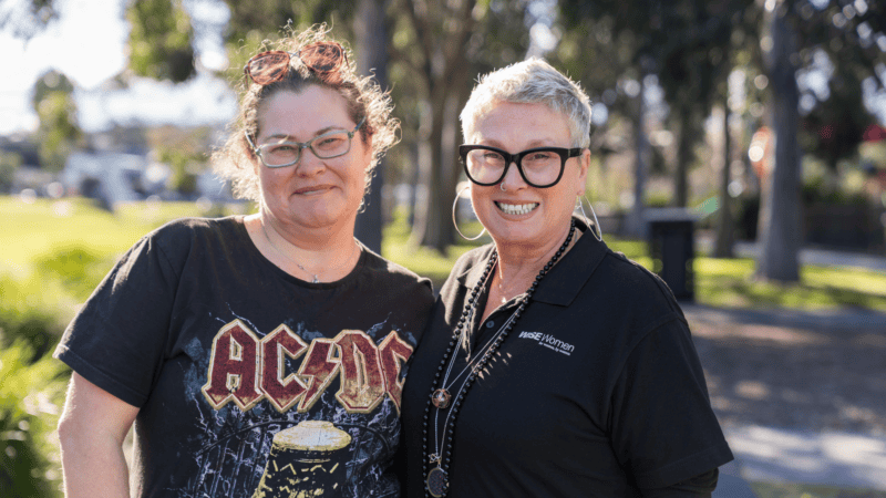 Two women stand together in a park, one in a black AC/DC t-shirt and the other in a black WISE Women polo shirt, both smiling at the camera.
