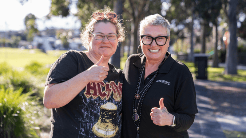 Two smiling women outdoors give a thumbs up, one wearing a black AC/DC t-shirt and the other a black WISE Women polo shirt.