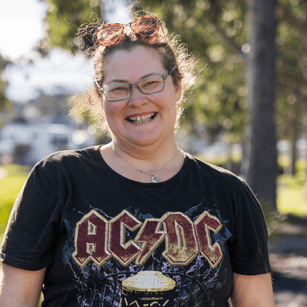 A smiling woman with brown hair and green-framed glasses stands in a sunny park, wearing a black AC/DC t-shirt.
