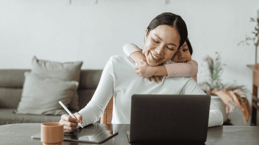 A woman smiles while working at a table with a laptop and tablet, as a young child hugs her from behind in a home setting.