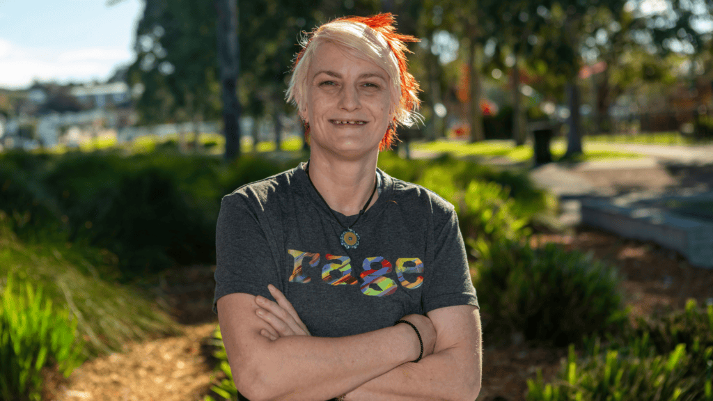 A smiling person with short blonde and red hair stands with arms crossed in a sunlit park, wearing a grey t-shirt with "rage" in rainbow text.