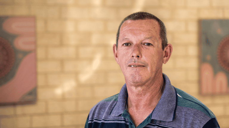 A man with short grey hair in a striped polo stands by a brick wall