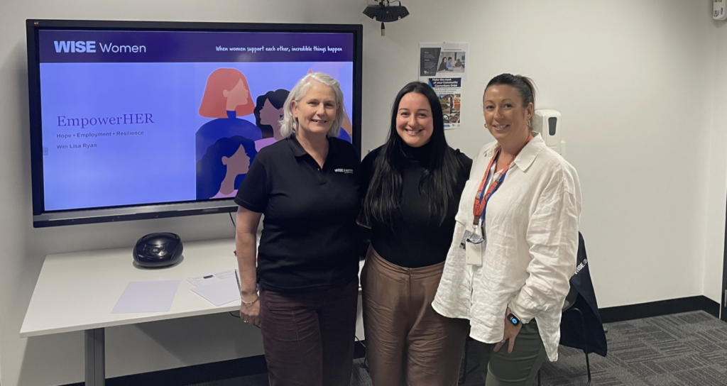 Three women stand together in front of a screen displaying the WISE Women EmpowerHER program in a meeting room.