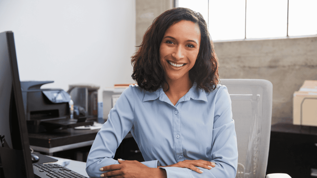 A smiling woman with dark hair sits at a desk in a modern office, wearing a light blue button-down shirt, with office equipment in the background.